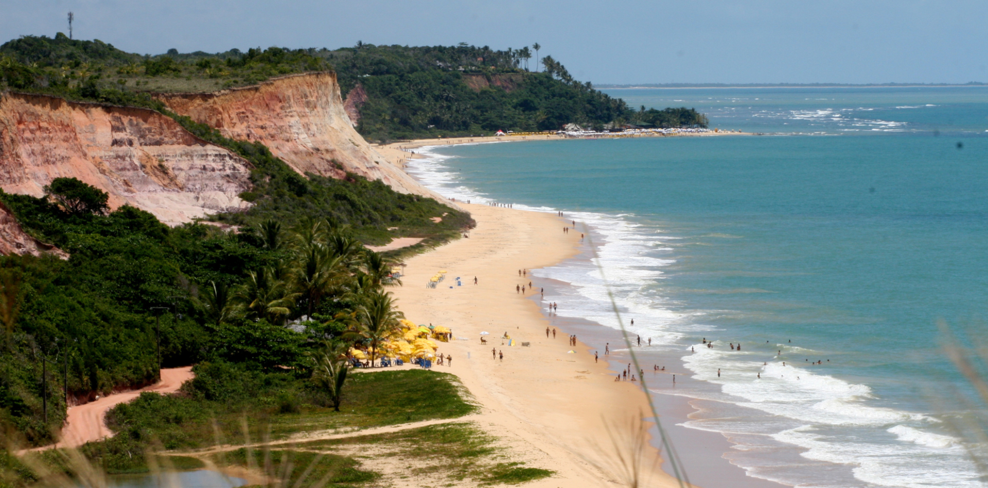 Praia com falesias em Arraial d Ajuda, vegetação entorno, mar azul e pessoas na praia