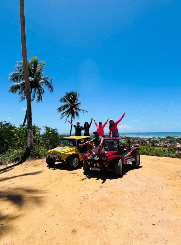 Grupo de quatro mulheres em cima de dois buggys estacionados na areia da praia em São Miguel dos Milagres, Alagoas. Elas estão com os braços levantados em comemoração, com coqueirais e o mar azul ao fundo.