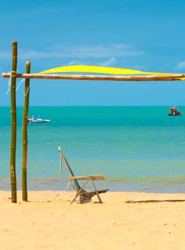 Uma tenda de praia montada com tecido amarelo vibrante, com cadeiras de praia embaixo para sombra. Ao fundo, a areia branca e o mar de São Miguel dos Milagres em tons de azul turquesa sob o sol.