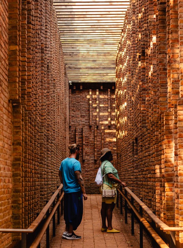 Fotografia de Bernardo Firme em ângulo zenital baixo, mostrando o corredor de entrada da Galeria Claudia Andujar no Instituto Inhotim. Duas pessoas observam atentas as paredes de tijolos aparentes, com a rampa do chão e o detalhe do teto compondo a cena em tons quentes e terrosos.