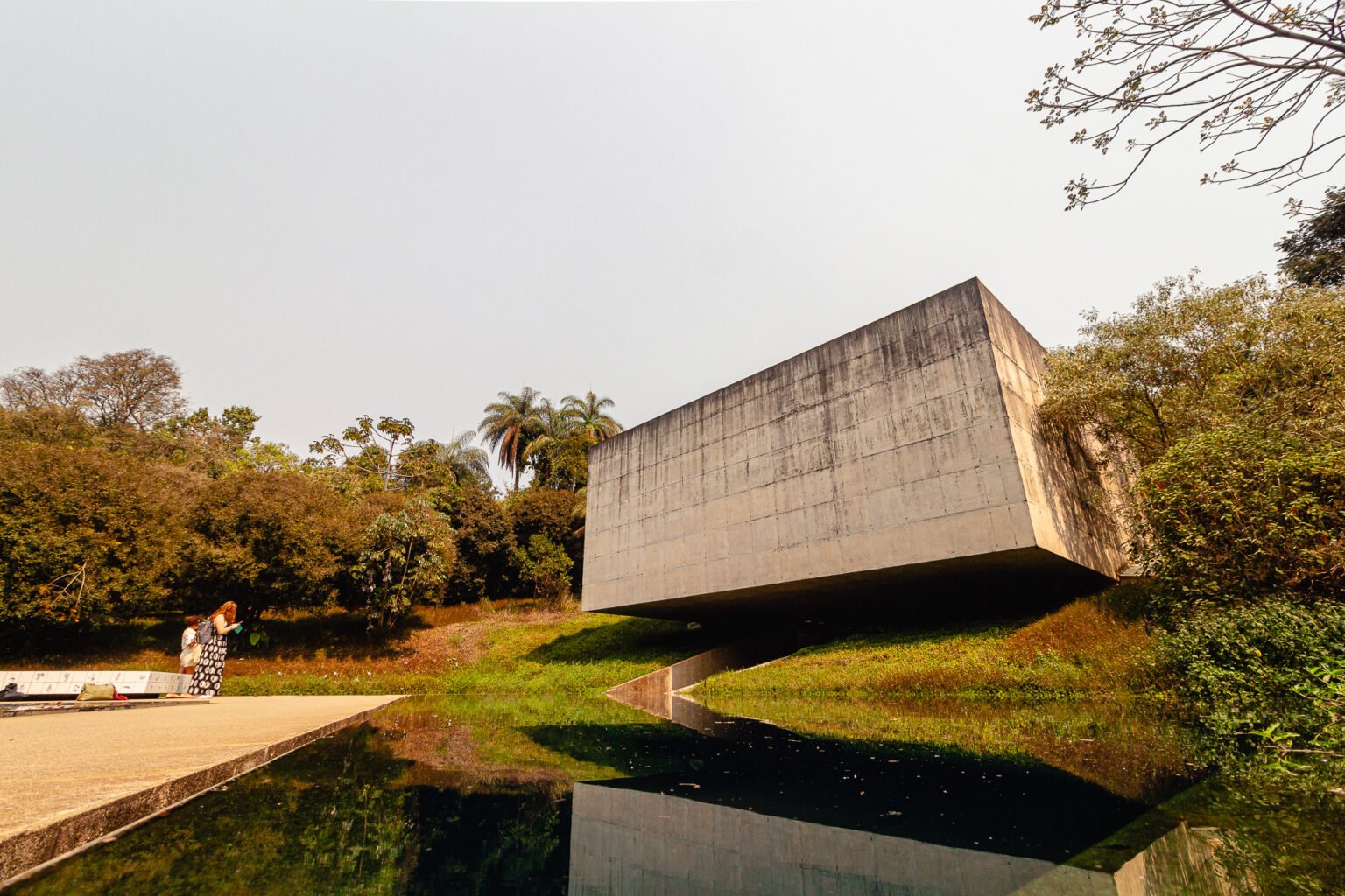 Fotografia panorâmica de Bernardo Firme mostrando a fachada de concreto da Galeria Adriana Varejão no Instituto Inhotim. Em primeiro plano, um espelho d'água reflete a estrutura e o céu, com duas pessoas em pé em frente à galeria, evidenciando a escala monumental da obra em meio ao paisagismo verde.