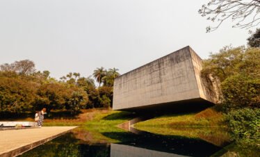 Fotografia panorâmica de Bernardo Firme mostrando a fachada de concreto da Galeria Adriana Varejão no Instituto Inhotim. Em primeiro plano, um espelho d'água reflete a estrutura e o céu, com duas pessoas em pé em frente à galeria, evidenciando a escala monumental da obra em meio ao paisagismo verde.