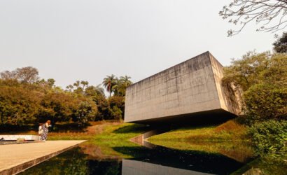 Fotografia panorâmica de Bernardo Firme mostrando a fachada de concreto da Galeria Adriana Varejão no Instituto Inhotim. Em primeiro plano, um espelho d'água reflete a estrutura e o céu, com duas pessoas em pé em frente à galeria, evidenciando a escala monumental da obra em meio ao paisagismo verde.