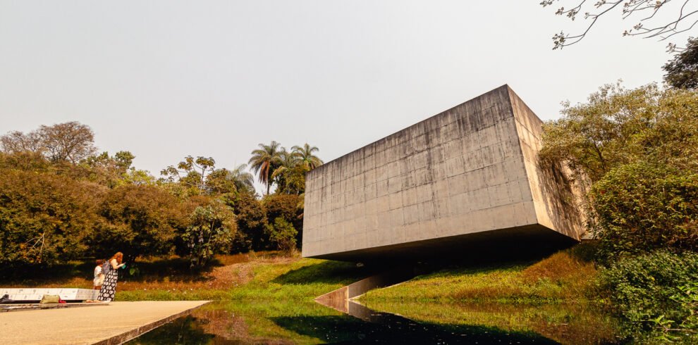 Fotografia panorâmica de Bernardo Firme mostrando a fachada de concreto da Galeria Adriana Varejão no Instituto Inhotim. Em primeiro plano, um espelho d'água reflete a estrutura e o céu, com duas pessoas em pé em frente à galeria, evidenciando a escala monumental da obra em meio ao paisagismo verde.