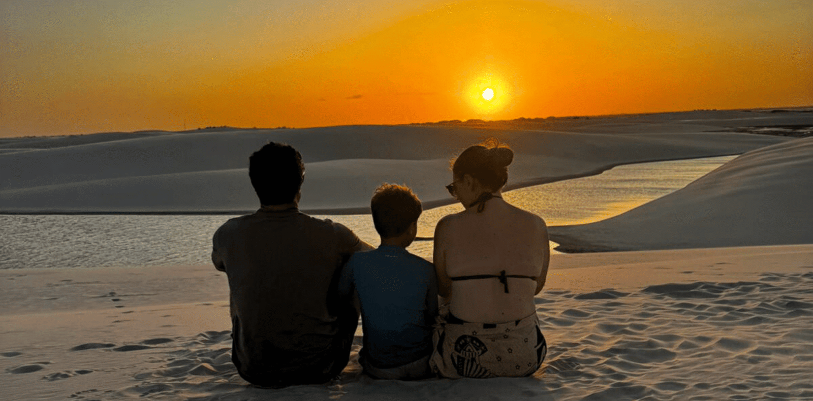 Pai, mãe e filho sentados lado a lado no topo de uma duna de areia branca nos Lençóis Maranhenses. Eles estão de costas, observando o pôr do sol alaranjado no horizonte vasto do parque nacional.