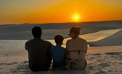 Pai, mãe e filho sentados lado a lado no topo de uma duna de areia branca nos Lençóis Maranhenses. Eles estão de costas, observando o pôr do sol alaranjado no horizonte vasto do parque nacional.