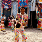 Uma criança de perfil, vestindo um traje típico regional colorido e ornamentado, realizando passos de uma dança tradicional do Maranhão em um pátio histórico. Ao fundo, detalhes da arquitetura local.
