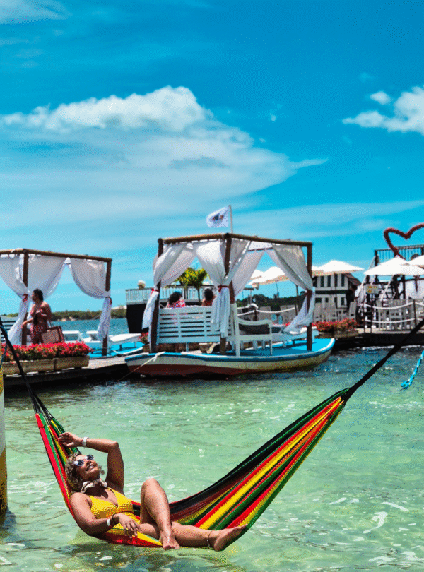 Mulher sorrindo deitada em uma rede dentro da água cristalina. Ela veste um maiô amarelo vibrante. Ao fundo, um deck de madeira com bangalôs rústicos de palha sobre a lagoa em Jericoacoara, Ceará.