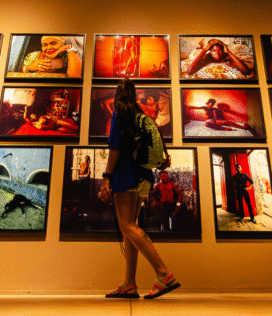 Fotografia de Bernardo Firme em ângulo contra-plongê (de baixo para cima), mostrando uma mulher de costas em Inhotim. Ela observa a exposição de Miguel Rio Branco na galeria homônima, com fotos de pessoas negras no Pelourinho, Salvador, em tons quentes e cenários simples de nudez cotidiana.
