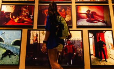 Fotografia de Bernardo Firme em ângulo contra-plongê (de baixo para cima), mostrando uma mulher de costas em Inhotim. Ela observa a exposição de Miguel Rio Branco na galeria homônima, com fotos de pessoas negras no Pelourinho, Salvador, em tons quentes e cenários simples de nudez cotidiana.