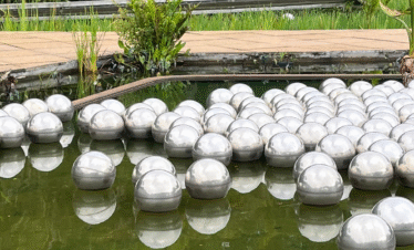 Fotografia de Bernardo Firme em Inhotim, mostrando a vista das passarelas de concreto que cruzam o espelho d'água da obra Narcissus Garden, de Yayoi Kusama. Centenas de esferas de aço inoxidável flutuam na superfície da água, refletindo o céu e a vegetação ao redor.
