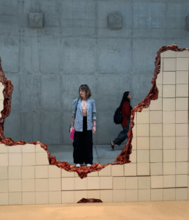 Fotografia do interior da Galeria Adriana Varejão em Inhotim. A imagem no formato vertical mostra a obra "Linda do Rosário", um muro de azulejos brancos rompido que revela órgãos e vísceras humanas esculpidas por dentro. Ao fundo, em uma parede cinza, uma mulher observa a obra por trás da fenda do muro.