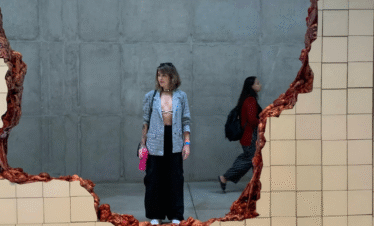 Fotografia do interior da Galeria Adriana Varejão em Inhotim. A imagem no formato vertical mostra a obra "Linda do Rosário", um muro de azulejos brancos rompido que revela órgãos e vísceras humanas esculpidas por dentro. Ao fundo, em uma parede cinza, uma mulher observa a obra por trás da fenda do muro.