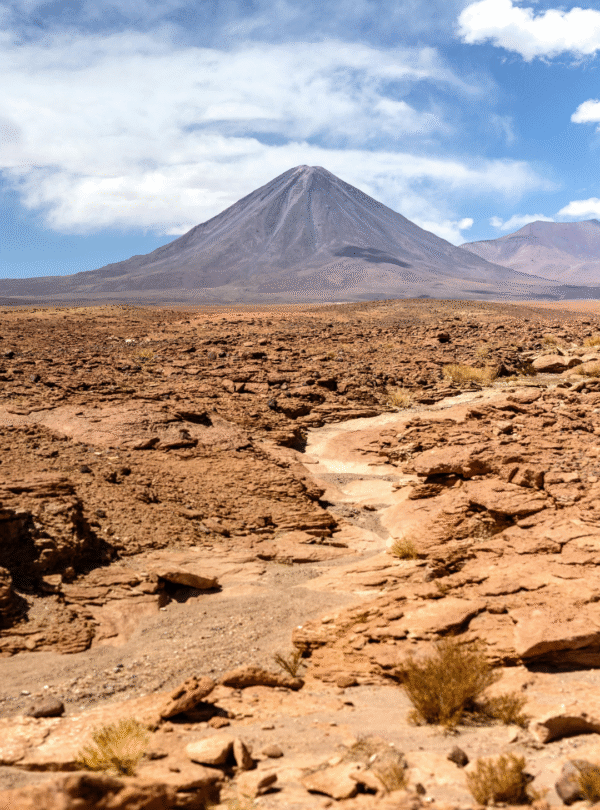 Fotografia panorâmica de uma estrada de terra que atravessa o Deserto do Atacama, com um vulcão ao fundo e nuvens brancas no céu azul.