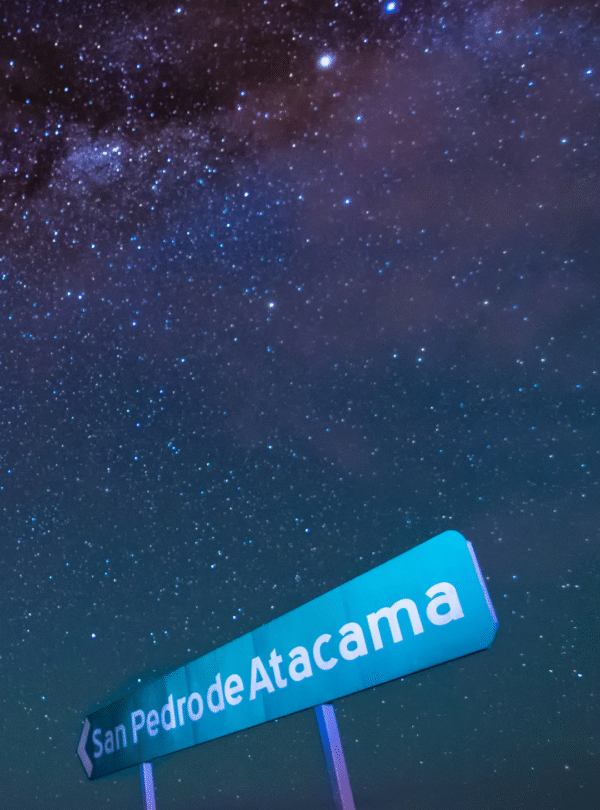 Fotografia de baixo para cima mostrando uma placa de sinalização escrita "San Pedro de Atacama" sob um céu noturno repleto de estrelas e a faixa da Via Láctea.