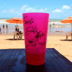 Copo rosa com arte do Espírito Santo sobre mesa de madeira, com praia, mar e guarda-sóis ao fundo em um dia ensolarado.